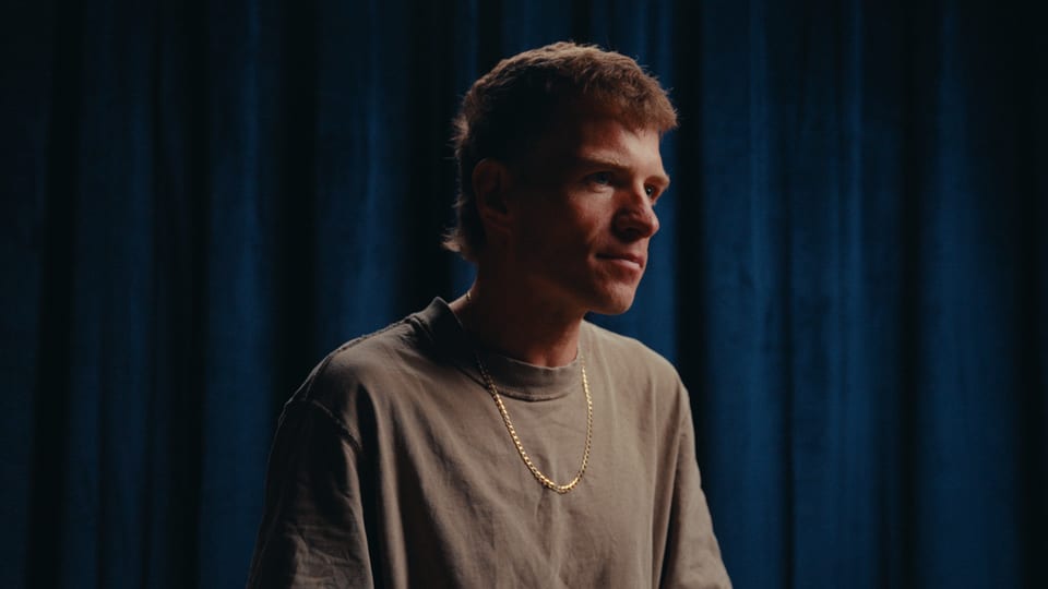 Athlete in a grey t-shirt and gold chain against a blue curtain