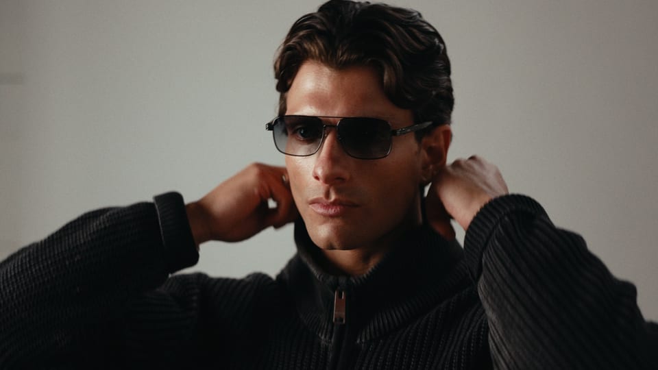 Man in sunglasses and a black knit jacket, hands at his collar