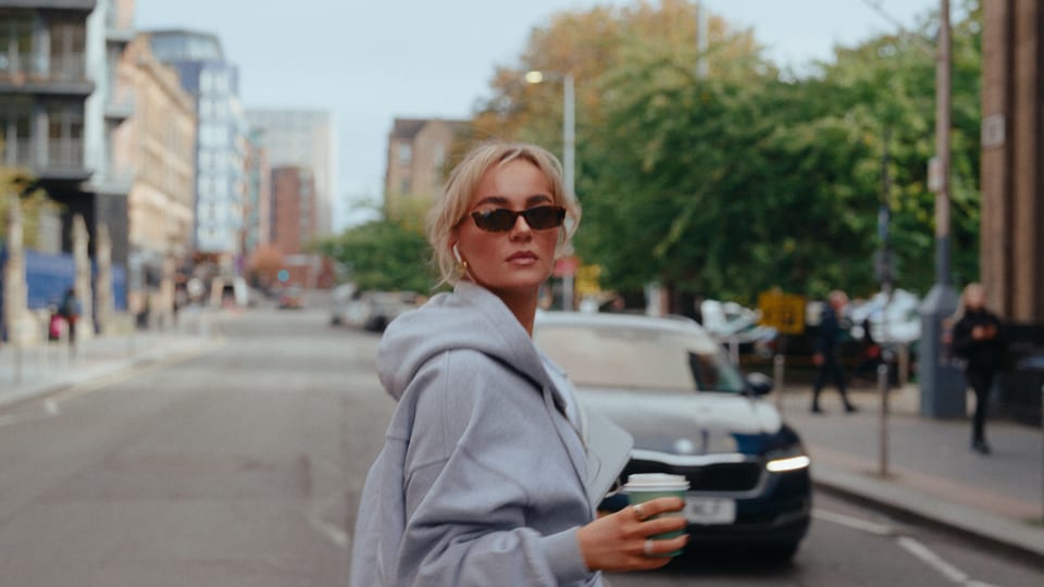 Woman in sunglasses and a grey hoodie with a coffee on a city street